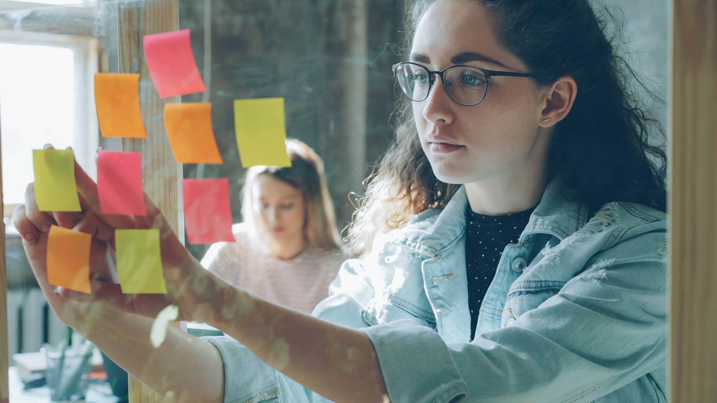 Woman placing colorful sticky notes on a glass wall while a colleague works in the background